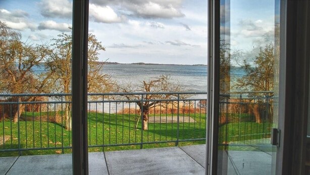 Blick vom Balkon mit Metallgeländer auf eine grüne Fläche mit Bäumen und ein Gewässer im Hintergrund.