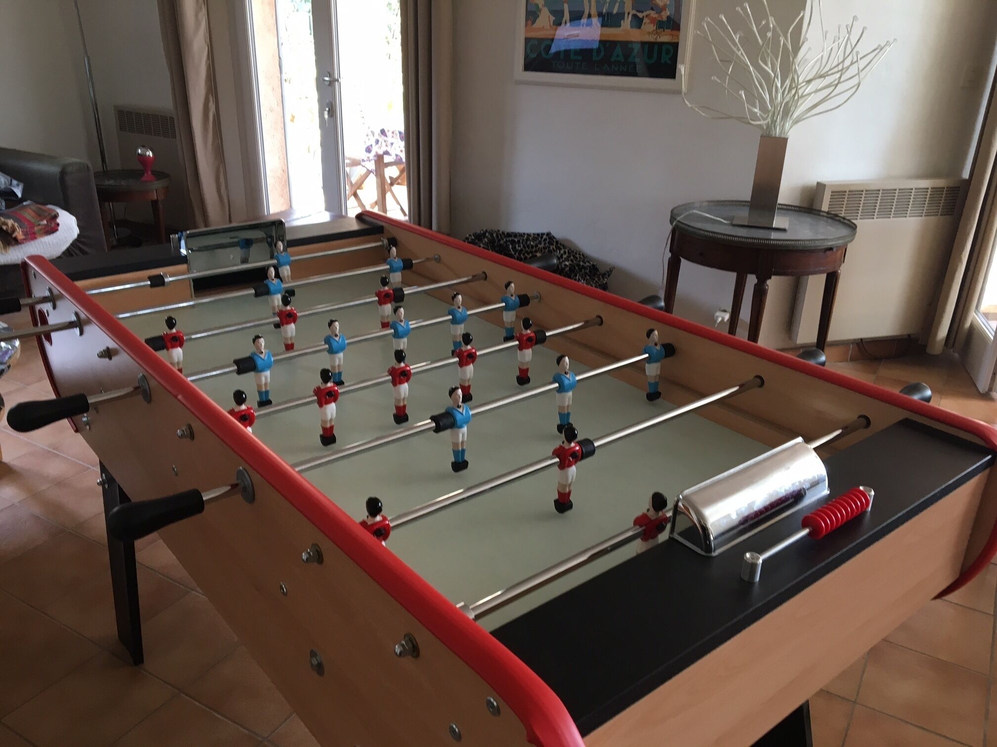 Una mesa de fútbol de mesa en una habitación con ventana, un radiador y una pequeña mesa con una planta.