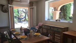 A dining area with a wooden table set for four, chairs, a window with a view of greenery and a statue, and a rustic cabinet. - Villa 6 People Saint Raphaël 5 min drive from the Beach, 2 min Golf courses (Saint-Raphaël)