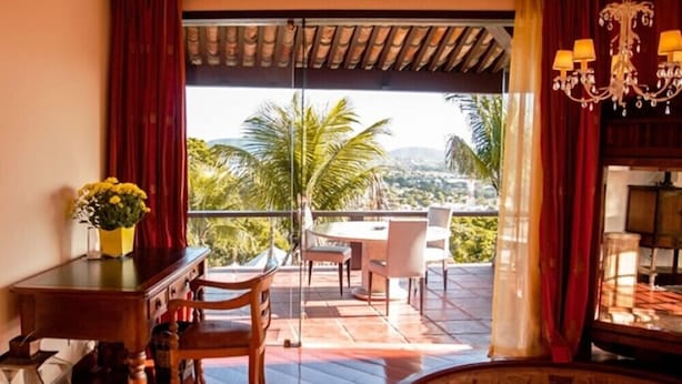 A room with a wooden table and chairs, a vase of flowers, and a view of palm trees and mountains through large windows. - Villa Felicche Búzios (Búzios)