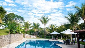 A swimming pool with lounge chairs and umbrellas, surrounded by palm trees and greenery. - Villa Felicche Búzios (Búzios)