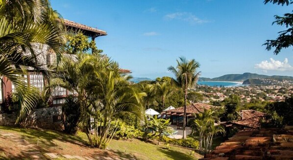 A coastal property with a stone house, lush greenery, and a view of the sea and distant hills.