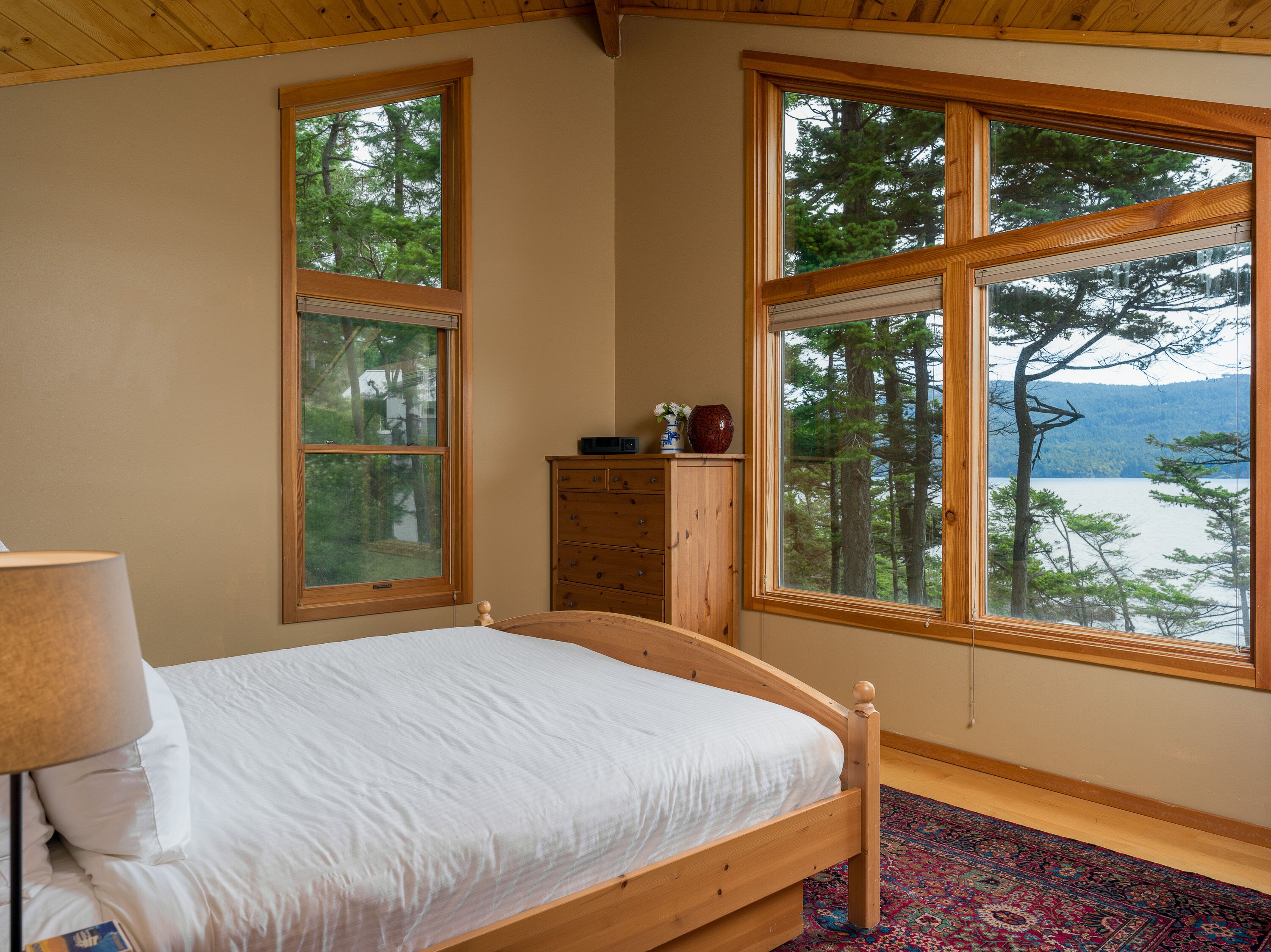 Ein Schlafzimmer mit einem hölzernen Bett, einem Schminktisch und großen Fenstern mit Blick auf Bäume und einen See.