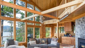 Amplia sala de estar con un sofá seccional gris, vigas de madera y ventanas grandes con vistas a un área boscosa.