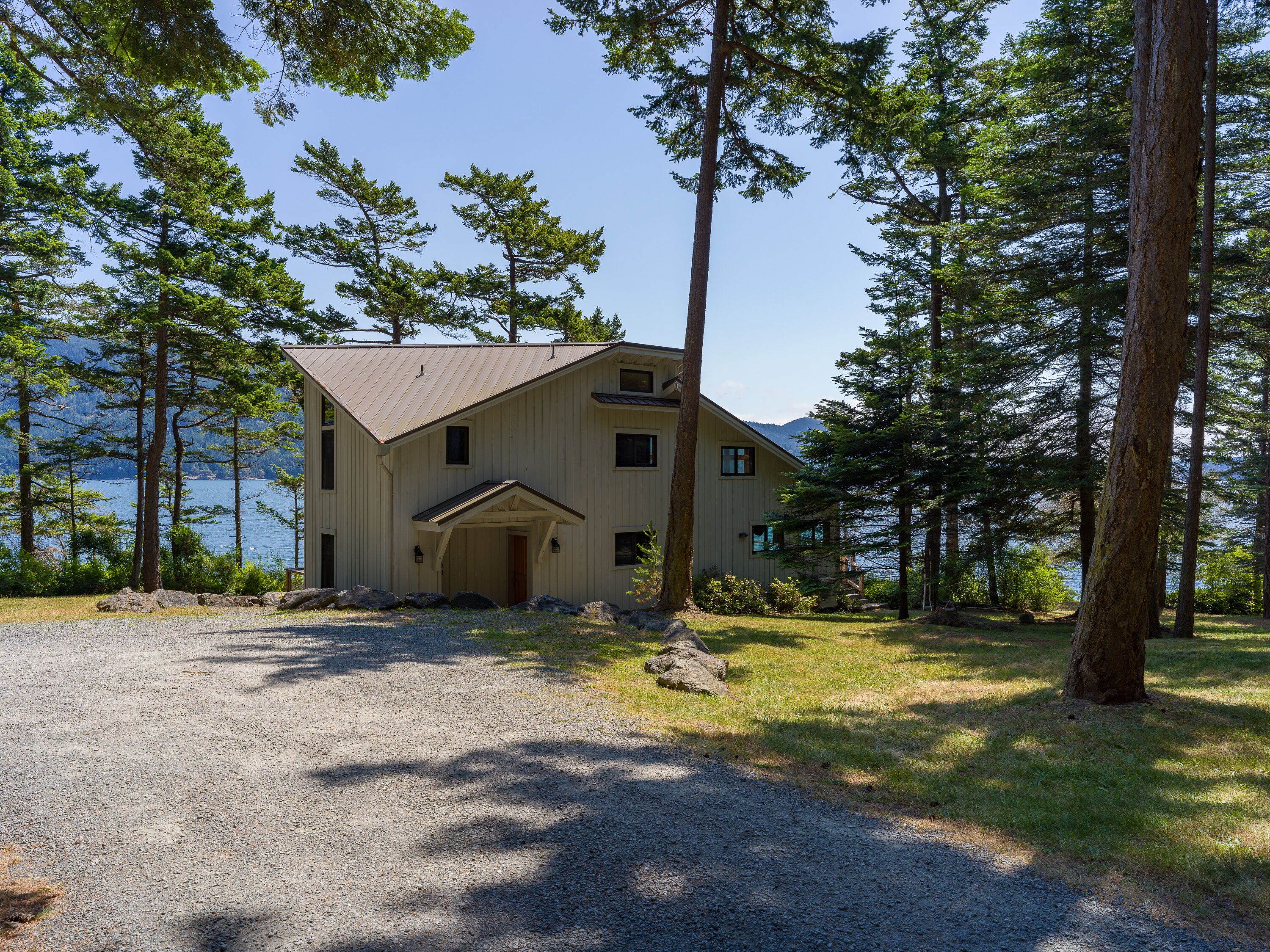 A two-story cabin with a gravel driveway, surrounded by tall trees and a lake view.
