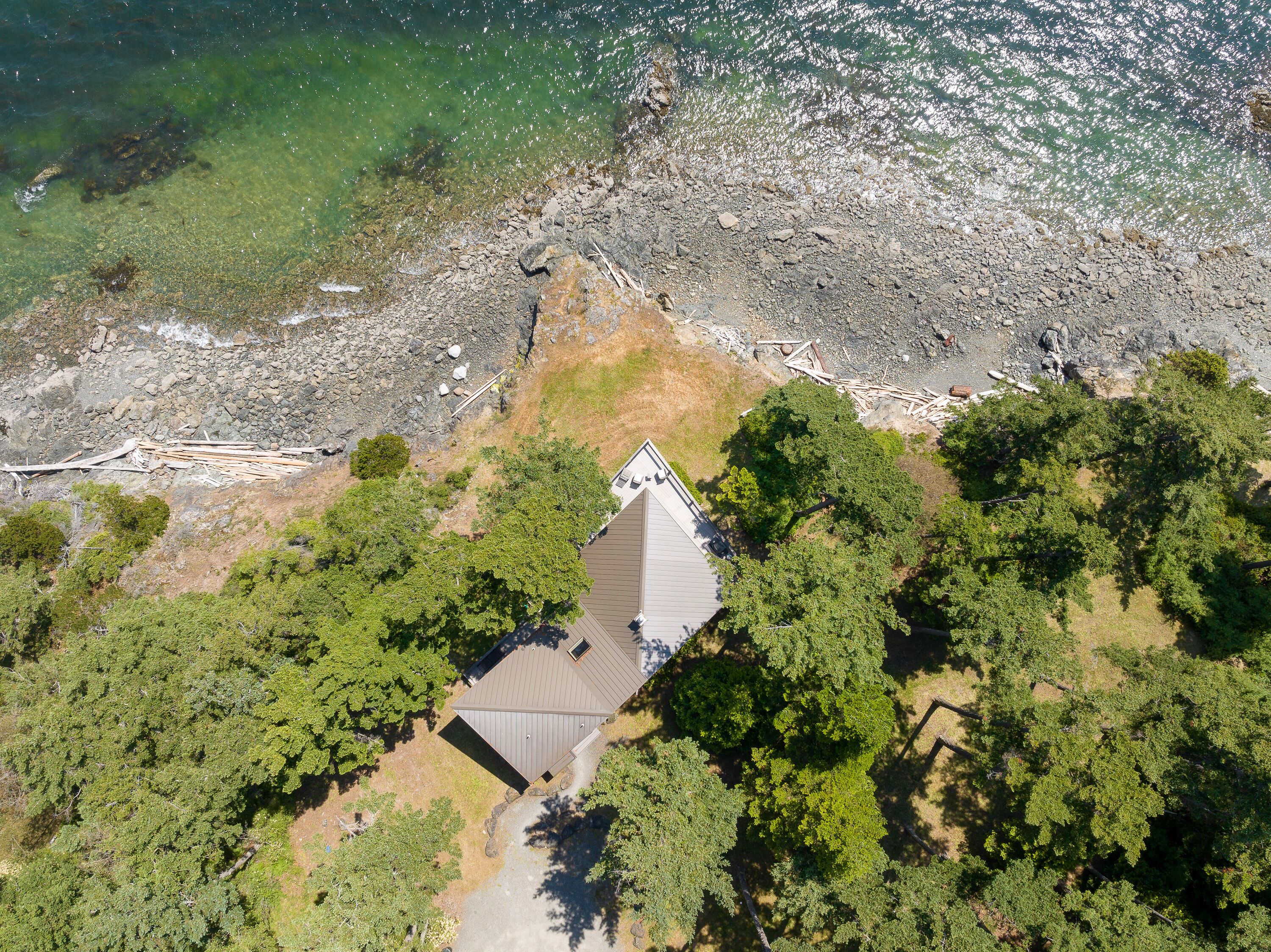 An aerial view of a house with a sloped roof, surrounded by dense green trees and a rocky shoreline.