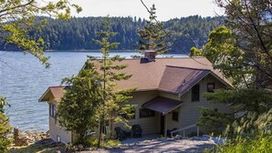 A house with a sloped roof, a chimney, and a porch with a chair. It is surrounded by trees and a body of water in the background. - Orcas Island, Juniper House #212 (Eastsound)