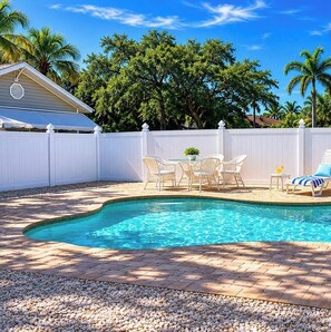 A swimming pool with a brick patio, white outdoor furniture, and a white fence.