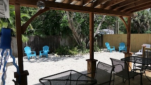 Eine überdachte Terrasse im Freien mit Metallstühlen und einem Tisch, umgeben von einer Holzdielenveranda und einer Sandfläche mit blauen Stühlen und einem Holzzaun.