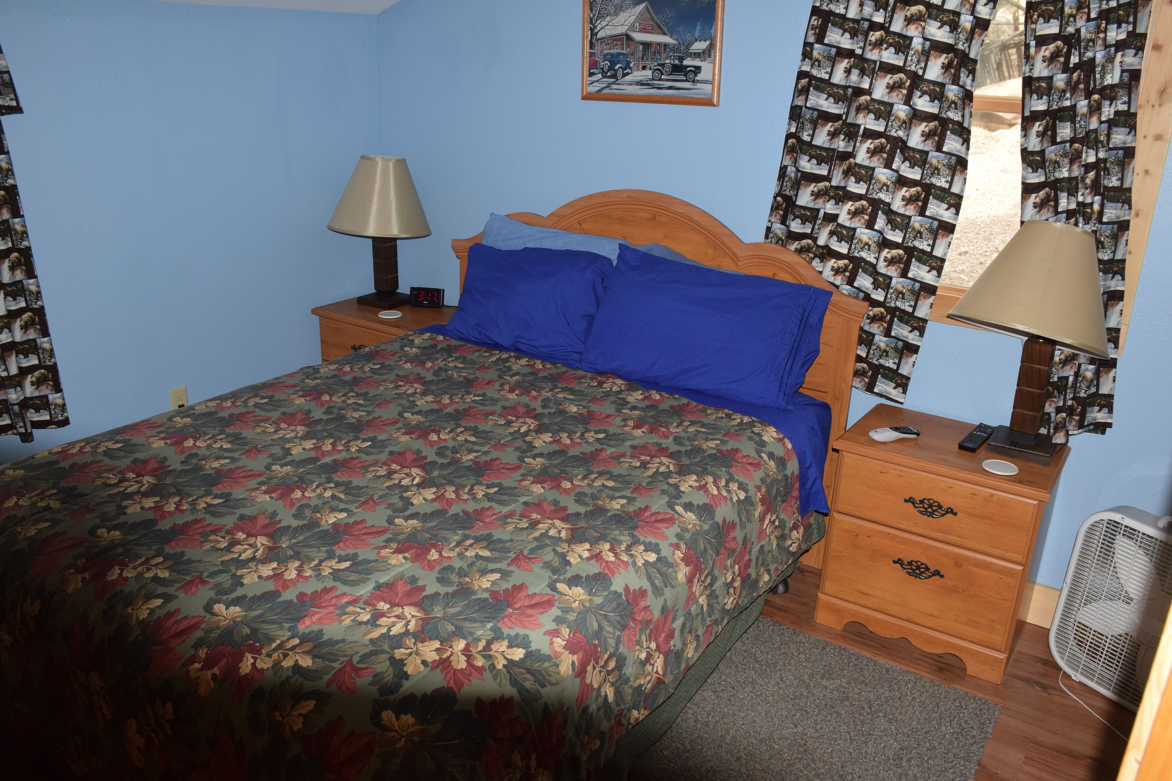 A bedroom with a floral bedspread, a wooden headboard, a nightstand with a lamp, a dresser with a mirror, and a wall-mounted picture.