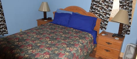 A bedroom with a floral bedspread, a wooden headboard, a nightstand with a lamp, a dresser with a mirror, and a wall-mounted picture.