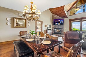 A dining area with a wooden table set for four, a chandelier, and a fireplace with a TV.