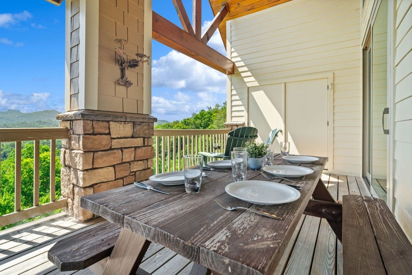An outdoor wooden deck with a table set for dining, a stone fireplace, and a view of the surrounding greenery.