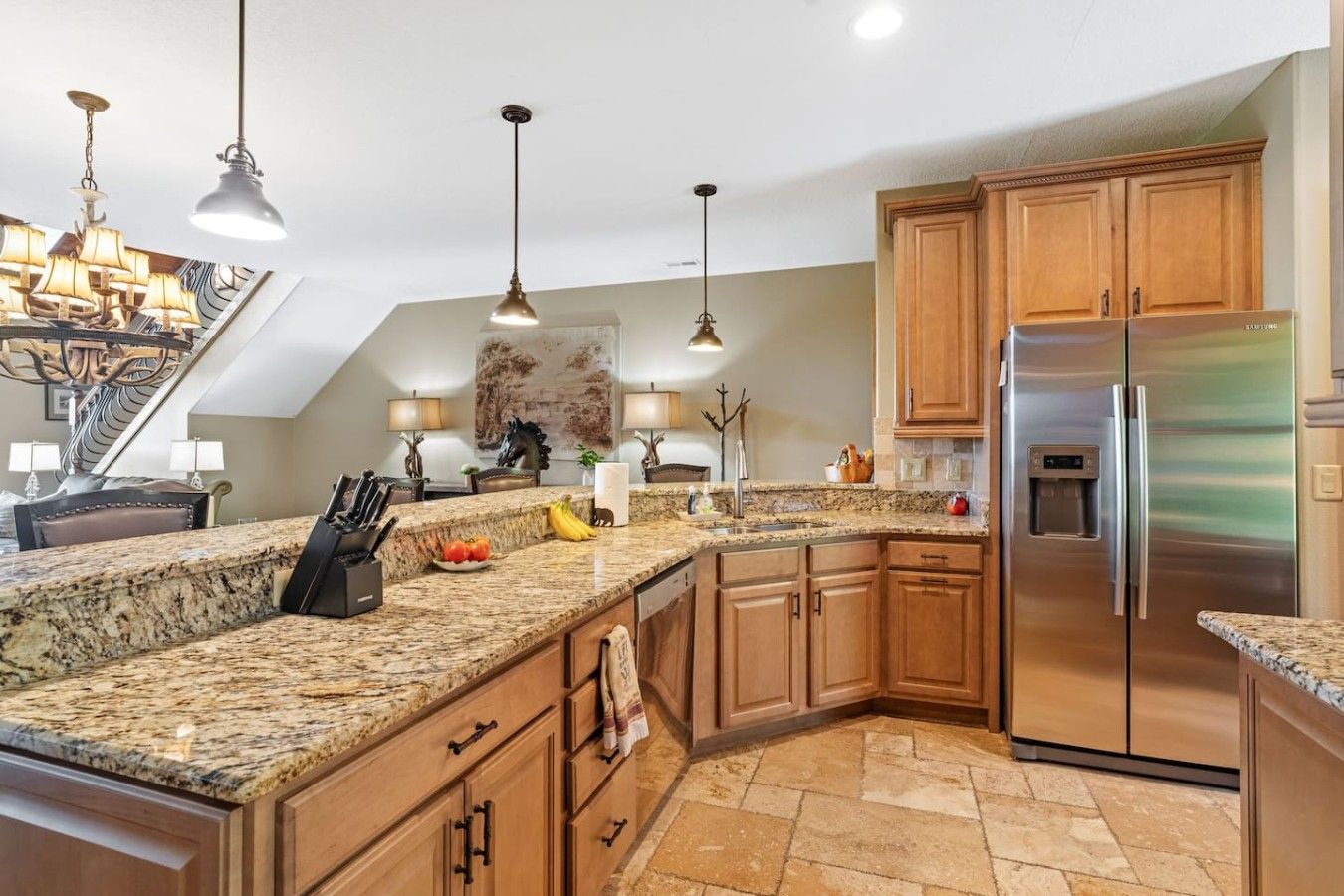 A kitchen with granite countertops, wooden cabinets, a stainless steel refrigerator, and pendant lighting.