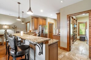 A kitchen with granite countertops, wooden cabinets, a stainless steel refrigerator, and a bar area with leather chairs.
