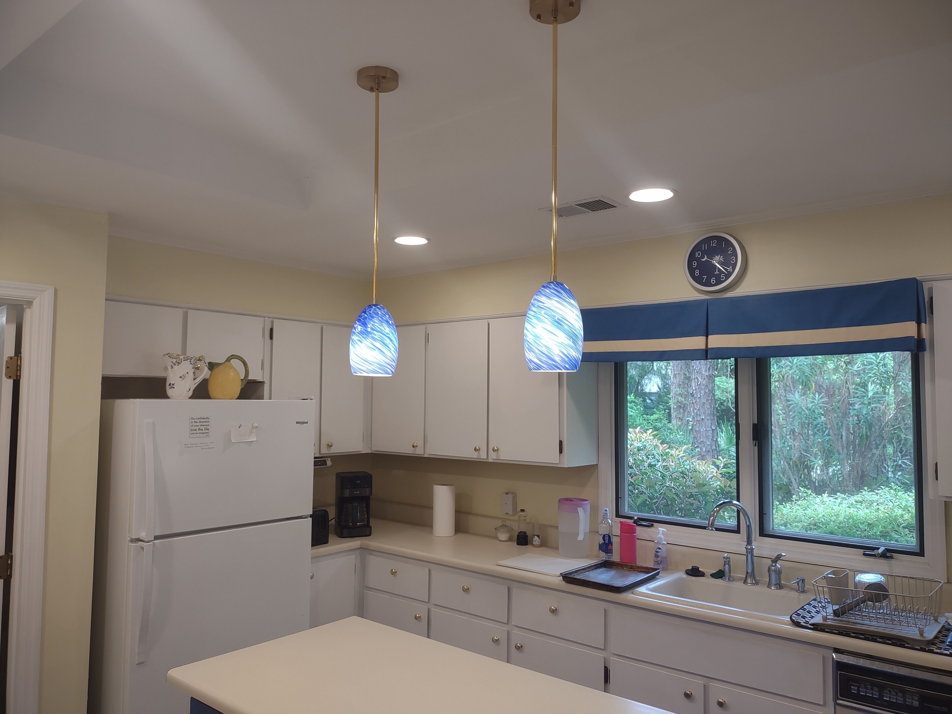A kitchen with white cabinets, a refrigerator, a coffee maker, a sink, and a window with a view of greenery.