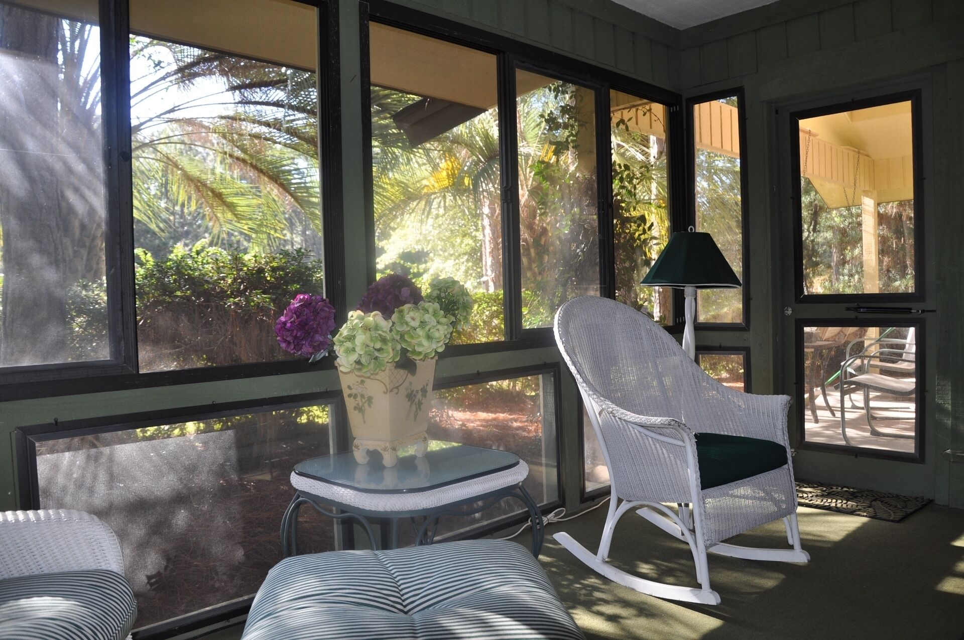 A sunlit room with large windows, a wicker chair, a small table, and a lamp.