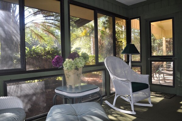 A sunlit room with large windows, a wicker chair, a small table, and a lamp.