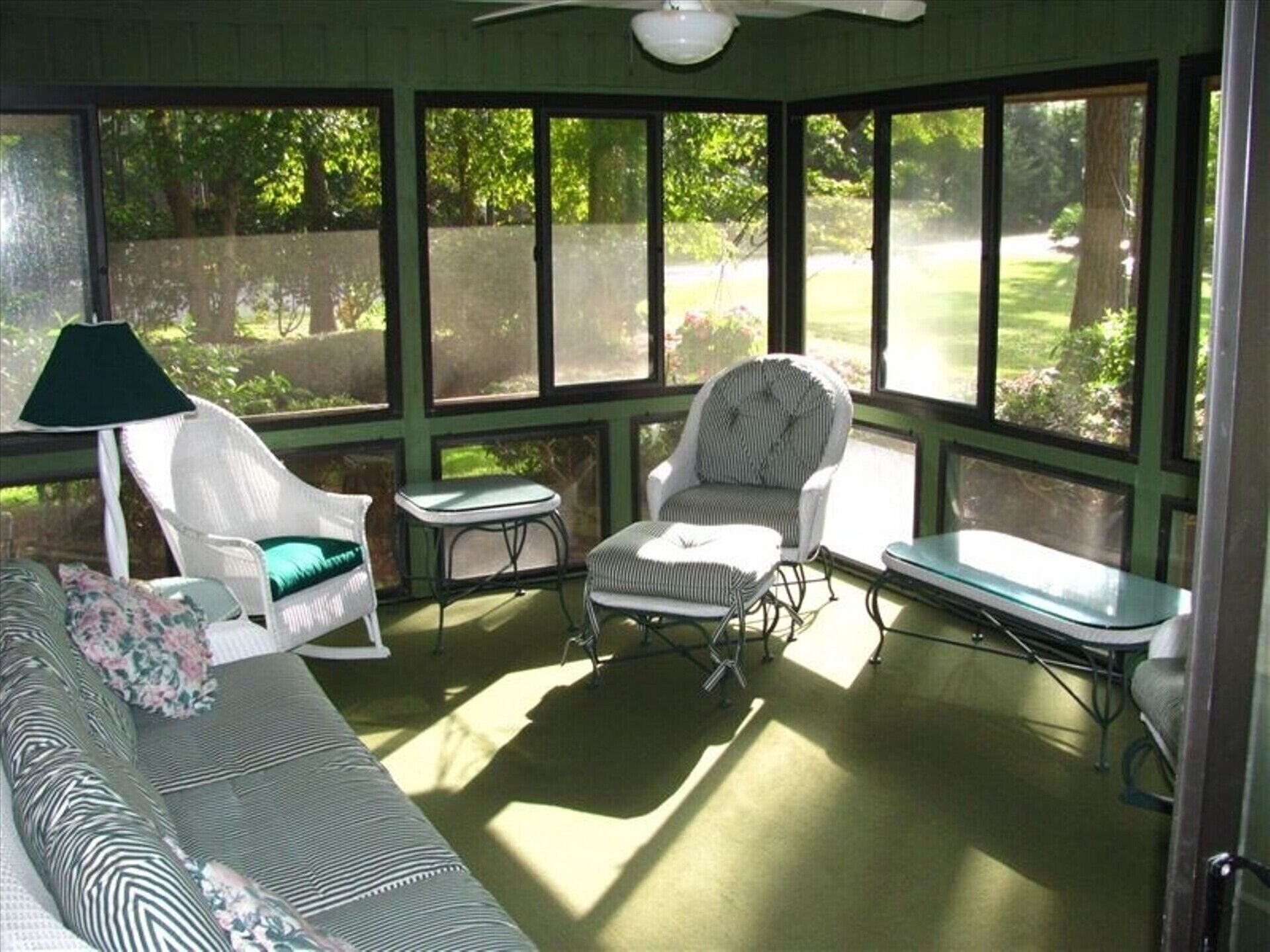 Ein Wintergarten mit grünem Boden, einem Deckenventilator und großen Fenstern. Zwei Sessel, ein kleiner Tisch und eine Couch mit gemusterten Kissen sind ebenfalls vorhanden.