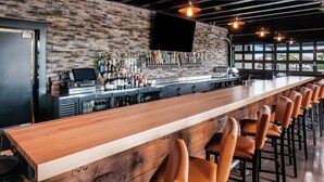 A bar area with a long wooden counter, multiple bar stools, a mounted TV, and a brick wall. - Cobblestone Hotel & Suites - Appleton International Airport (Neenah)