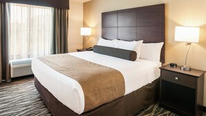A hotel room with a large bed, two bedside tables with lamps, a window with curtains, and a wall-mounted air conditioner. - Cobblestone Hotel & Suites - Appleton International Airport (Neenah)