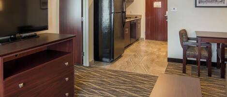 A compact hotel room with a TV, a refrigerator, a dining table with chairs, and a patterned rug.