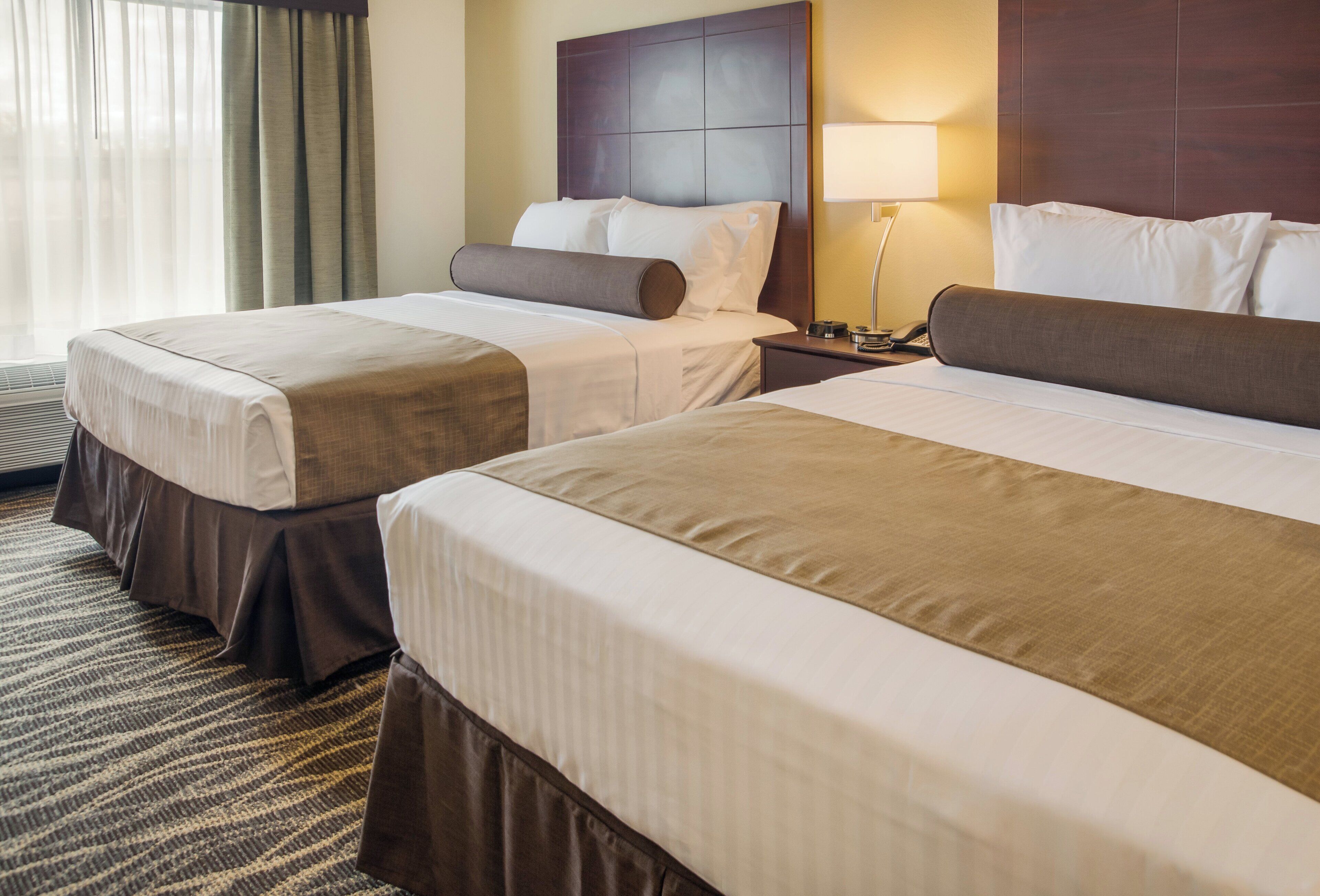 A hotel room with two beds, a nightstand, a lamp, and a window with curtains.