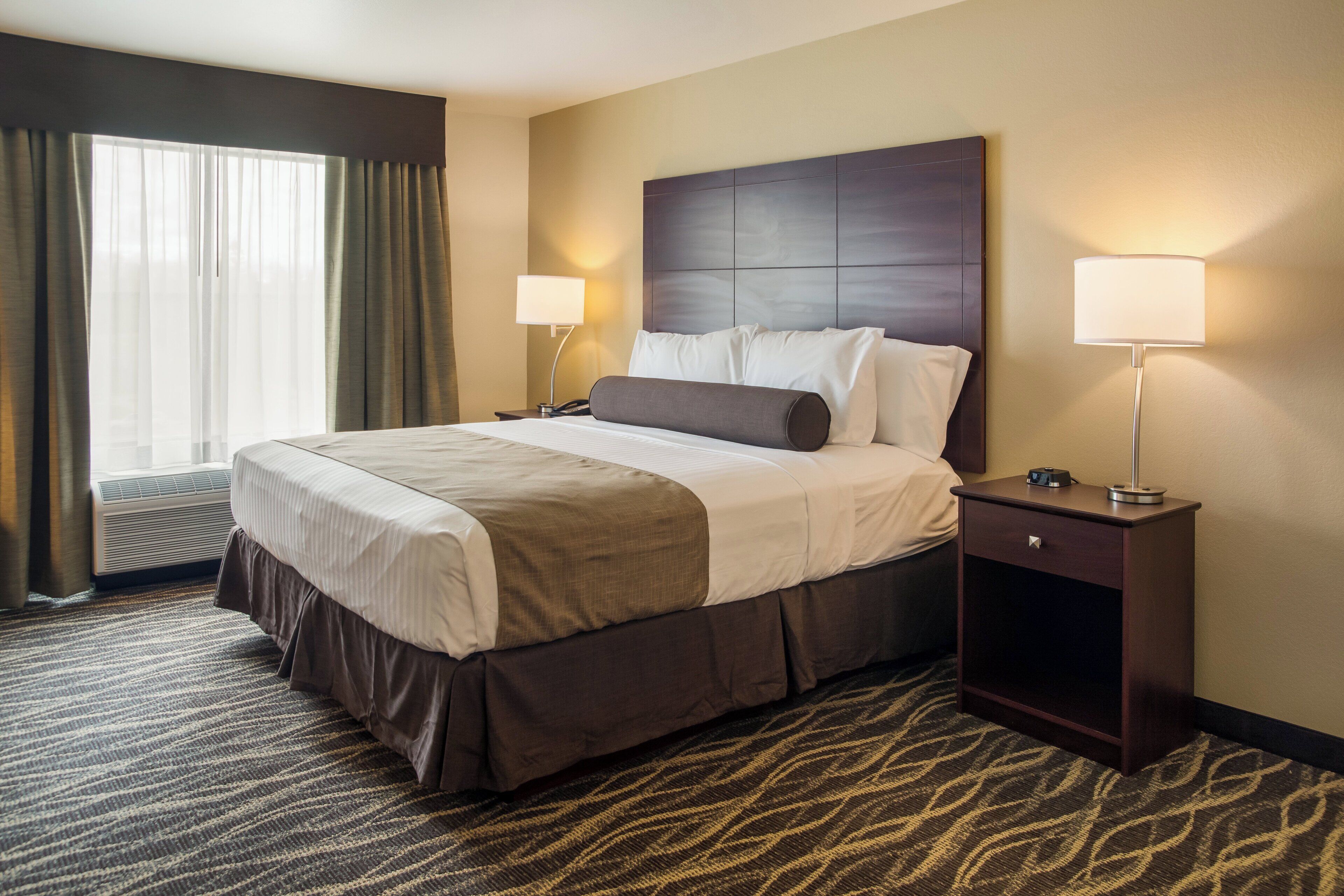 A hotel room with a large bed, two bedside tables, a window with curtains, and a patterned carpet.