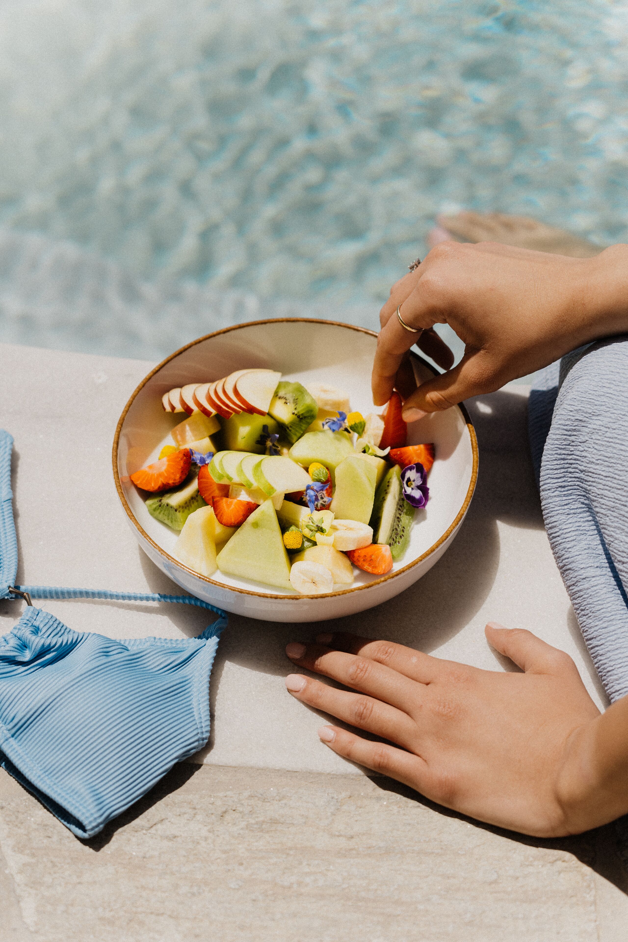 A bowl of fruit salad with a variety of fruits including apples, oranges, and kiwi, placed on a table near a pool.