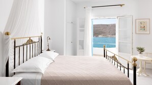 A bedroom with a bed, a bedside table, a lamp, a window with a view of the sea, and a door leading to another room. - Melian Boutique Hotel & Spa (Milos)
