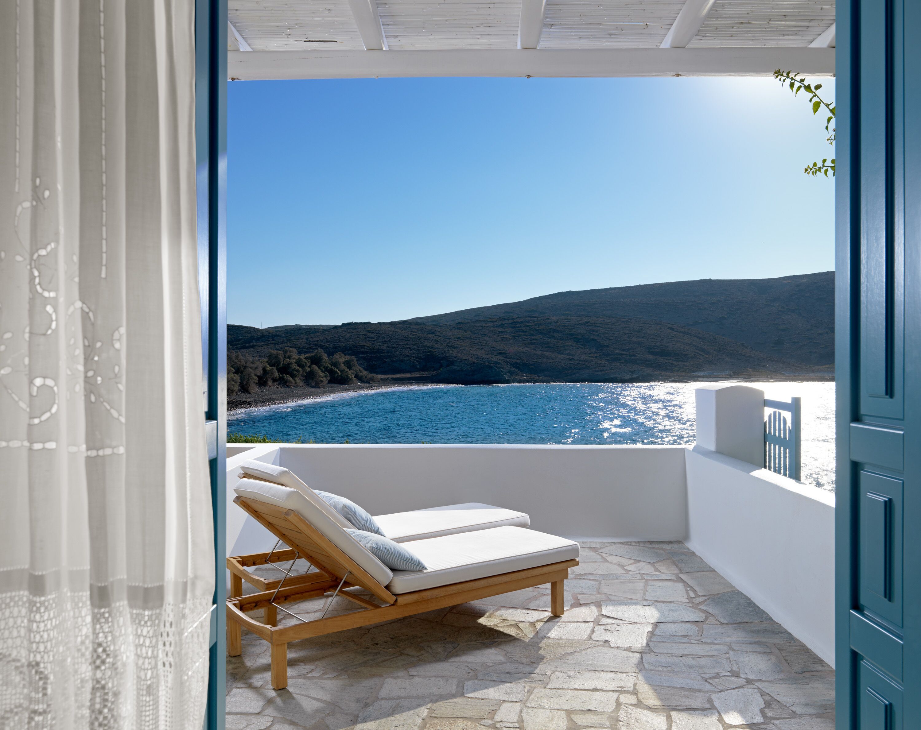 A balcony with a stone floor, a wooden lounge chair, and a view of the sea and hills.