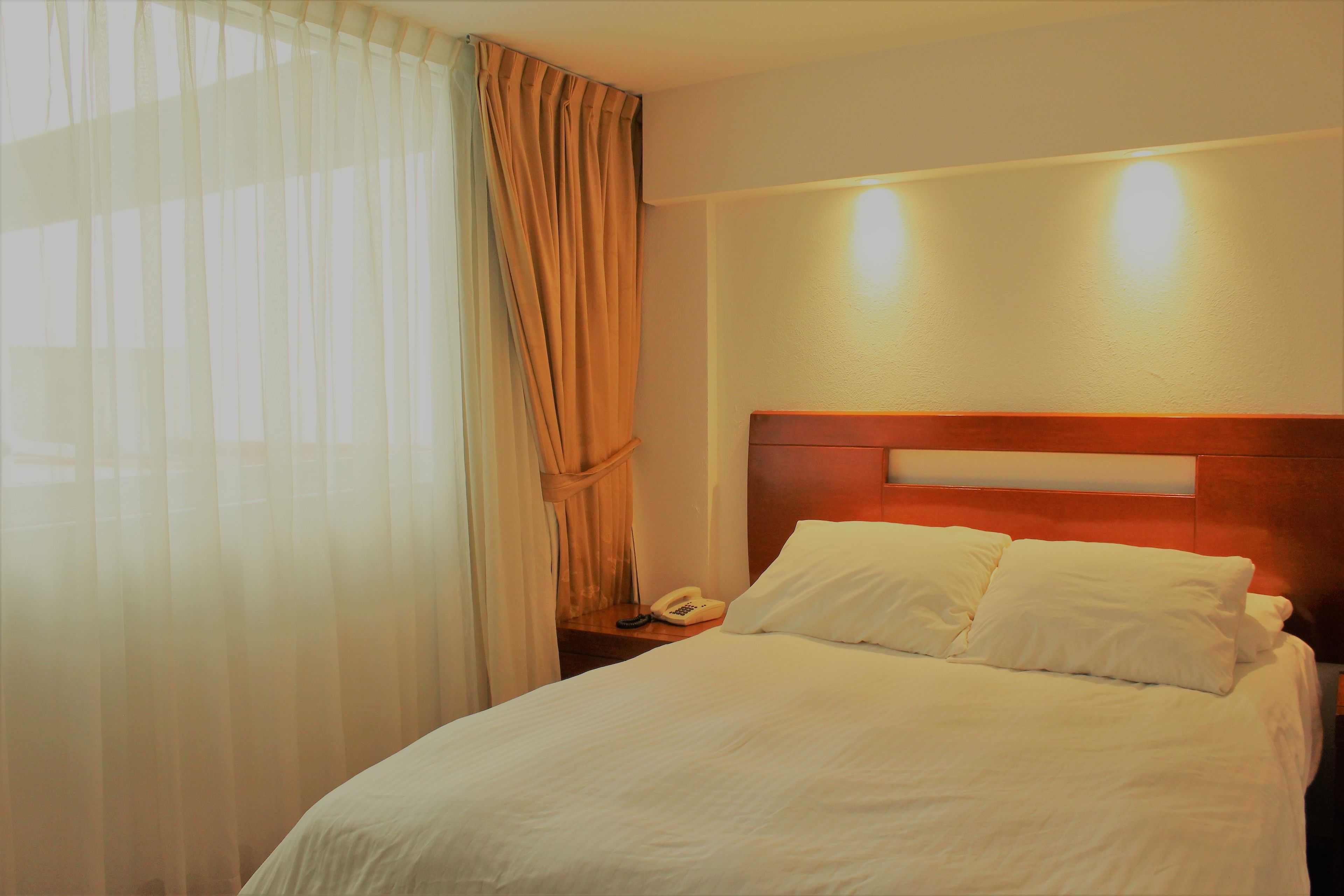 Ein Hotelzimmer mit einem ordentlich bezogenem Bett, einem Nachttischtelefon und einem zur Seite zurückgezogenen Vorhang.