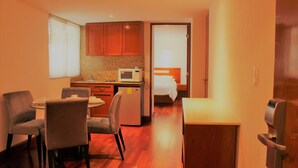 Habitación de hotel compacta con kitchenette, zona de comedor con mesa redonda y sillas, cama con cabecera blanca y puerta con manija.