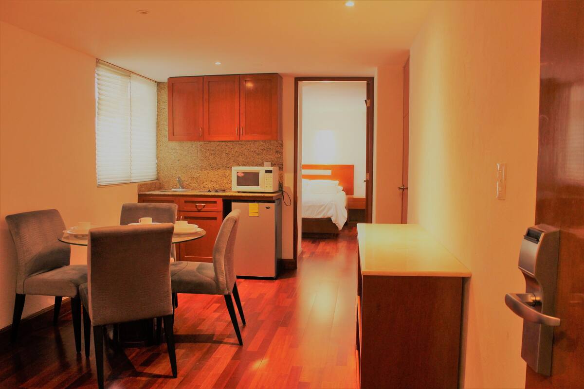 Habitación de hotel compacta con kitchenette, zona de comedor con mesa redonda y sillas, cama con cabecera blanca y puerta con manija.