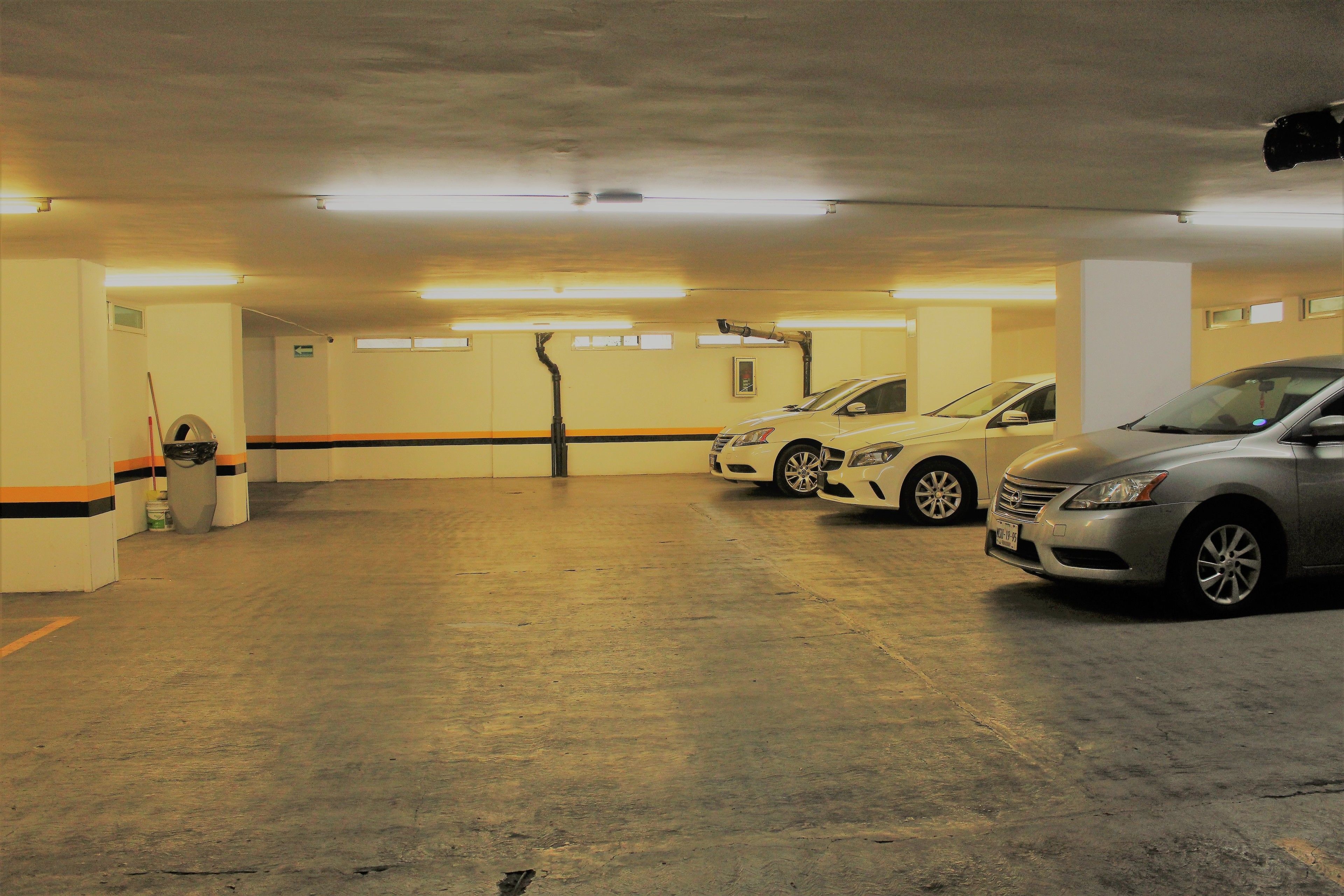 A parking garage with three cars parked in a row, fluorescent lighting, and a concrete floor.