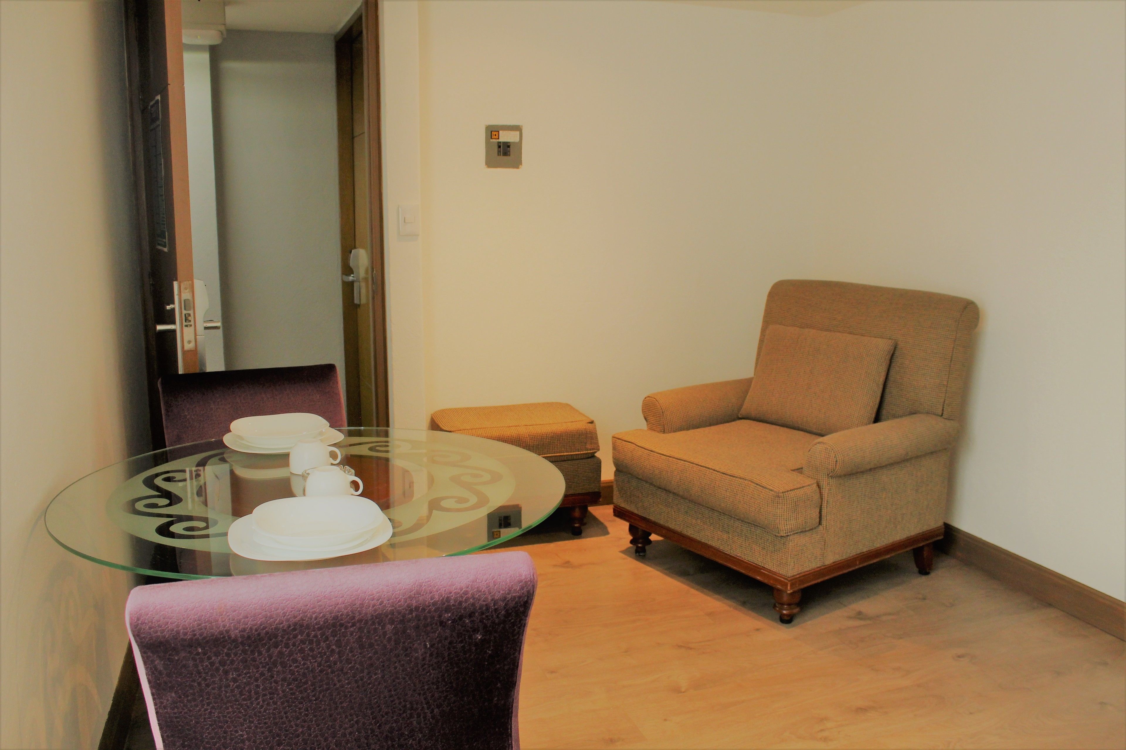 A small room with a glass table, a purple chair, a beige armchair, and a wooden floor.