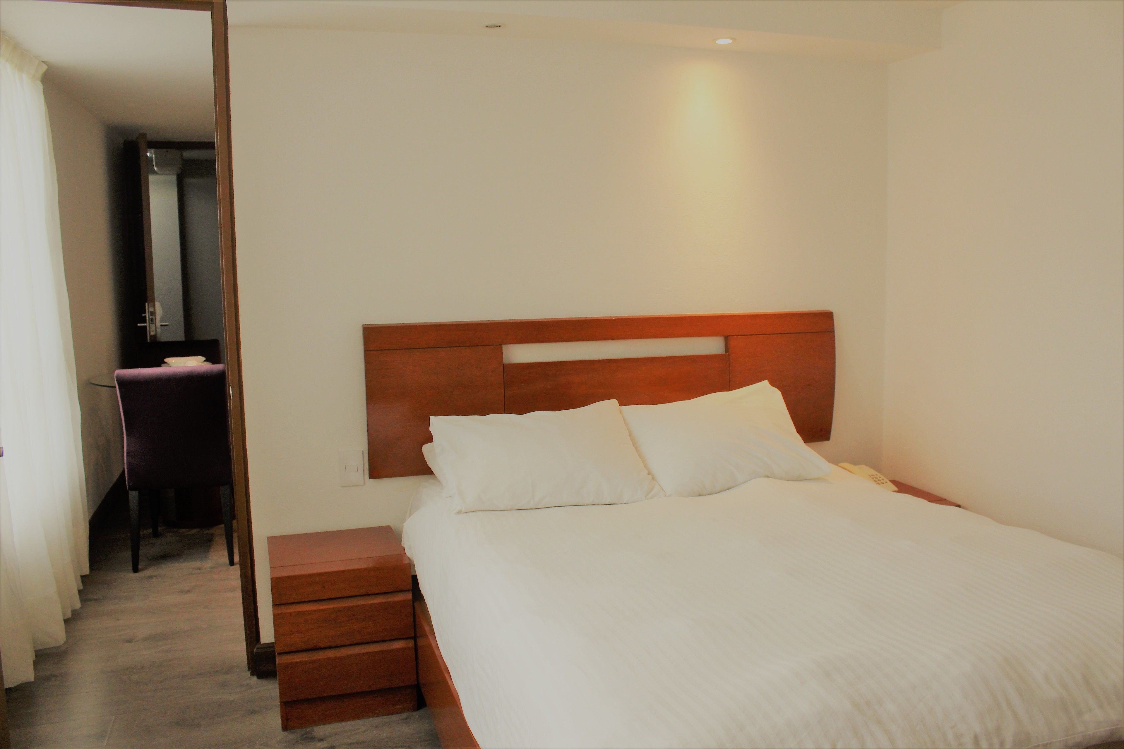 Una cama bien tendida con cabecera de madera, una mesita de noche y una silla en una habitación con paredes blancas y suelo de madera.