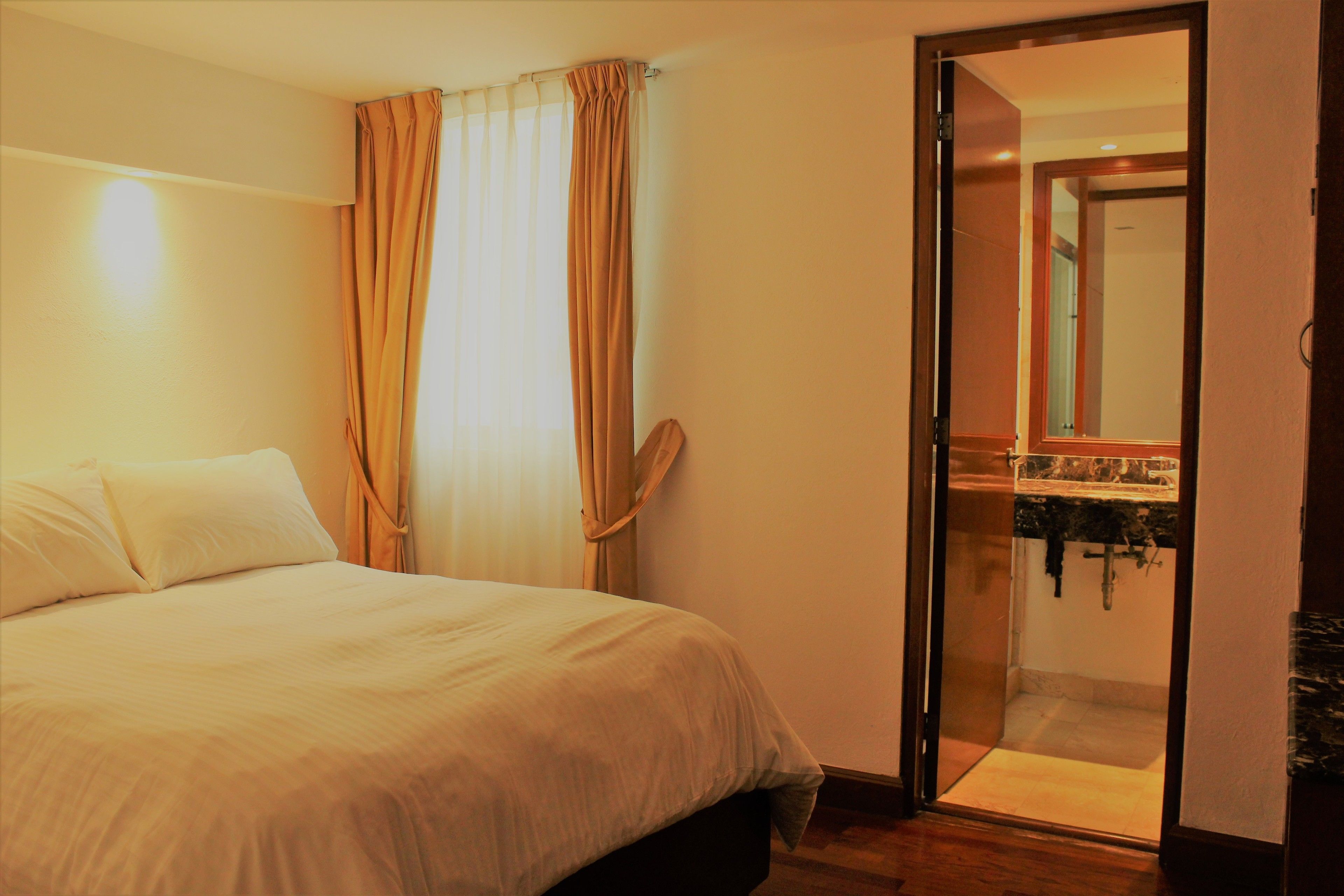 Habitación de hotel con una cama, un baño visible a través de una puerta de vidrio y una zona de ducha.