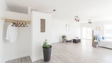 A spacious living room with a wooden floor, a black trash bin, a wooden coat rack with clothes, a potted plant, and a bed with a white headboard.