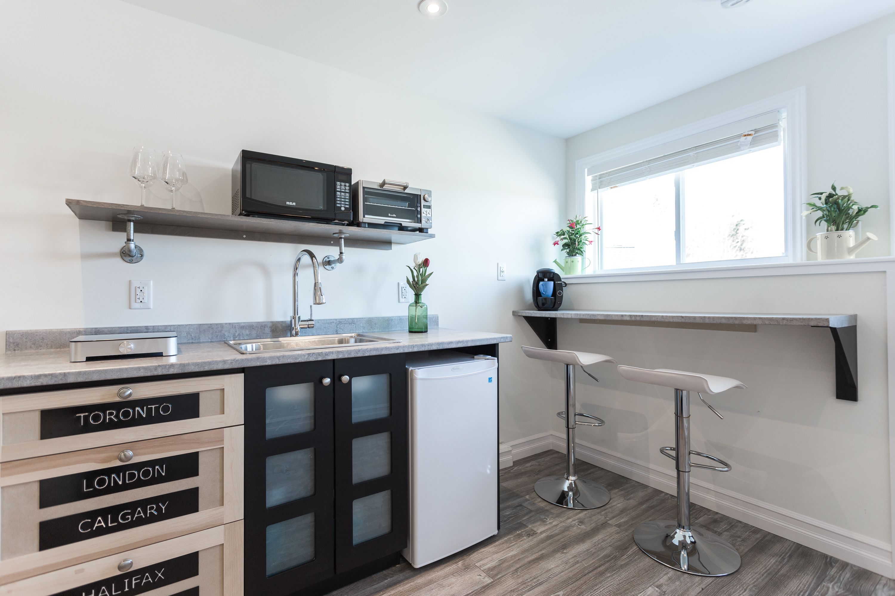 A modern kitchen with a built-in microwave, a refrigerator, and a sink. There are bar stools and a window with plants on the windowsill.