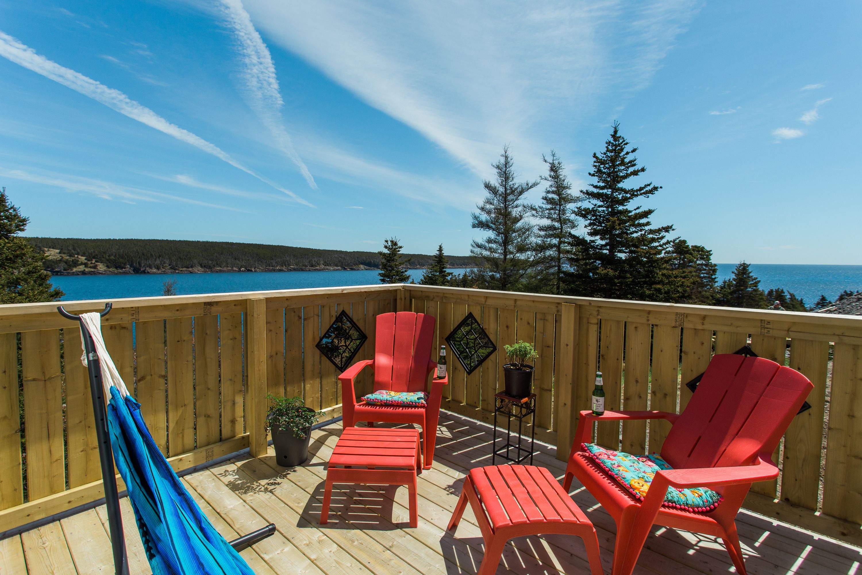 A wooden deck with red Adirondack chairs, a hammock, and potted plants.