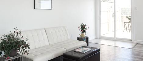 Una sala moderna con un sofá blanco, un reposapiés negro y una estantería. Hay un ventanal corredero que da a un patio exterior.