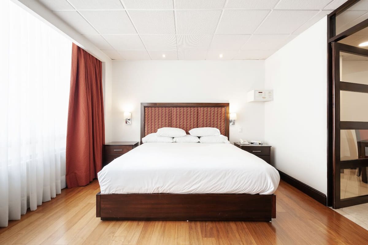 Una habitación de hotel moderna con piso de madera, una cama grande con ropa blanca, un cabecero estampado, lámparas de noche y una puerta corrediza de vidrio.