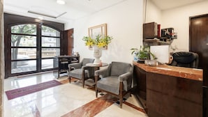 A reception area with a marble floor, a reception desk, two grey armchairs, a vase with yellow flowers, and a large window with a view of buildings and trees.