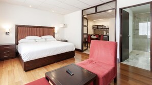 A hotel room with a bed, a red chair, a small table, a remote control, and a bathroom with a shower and toilet.