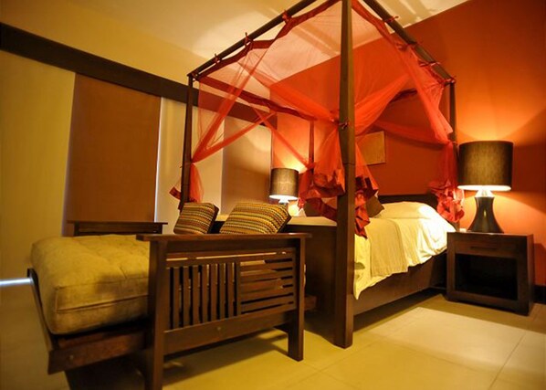 A four-poster canopy bed with a wooden frame and red fabric canopy, flanked by two bedside tables with lamps, a wooden bench, and a bed with white bedding.
