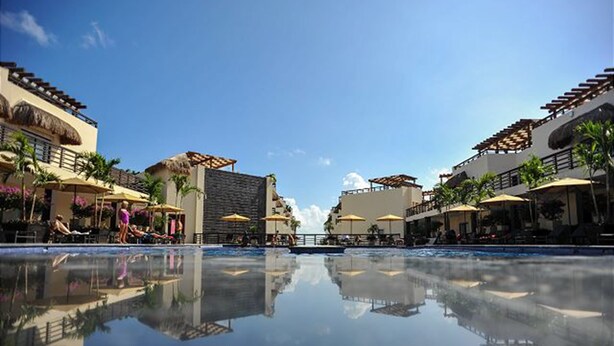 A resort pool with a clear reflection, surrounded by palm trees and umbrellas, and buildings with balconies. - Aldea Thai 35 by Tripintravel (Playa del Carmen)