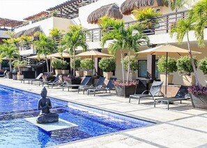 A pool area with lounge chairs and umbrellas, a statue in the water, and a building with a thatched roof.