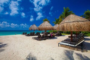 Ein Strand mit Reetdachschirmen, Liegestühlen und Palmen.