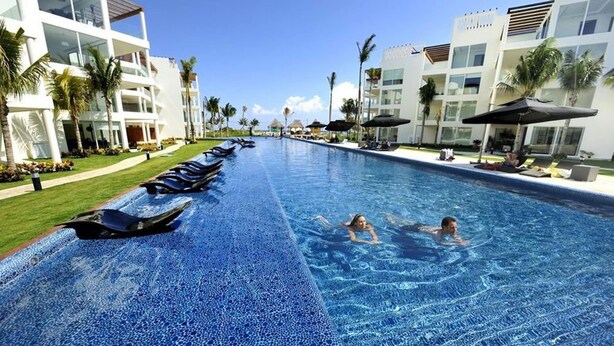 A swimming pool with lounge chairs and umbrellas, surrounded by modern buildings and palm trees. - Elements GH18 by Tripintravel (Playa del Carmen)